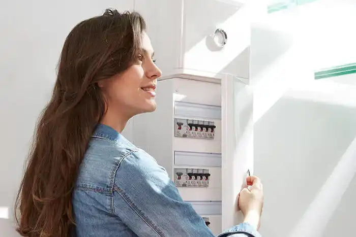 Femme souriante en veste en jean ouvrant un tableau électrique blanc dans un escalier, rénovation électrique