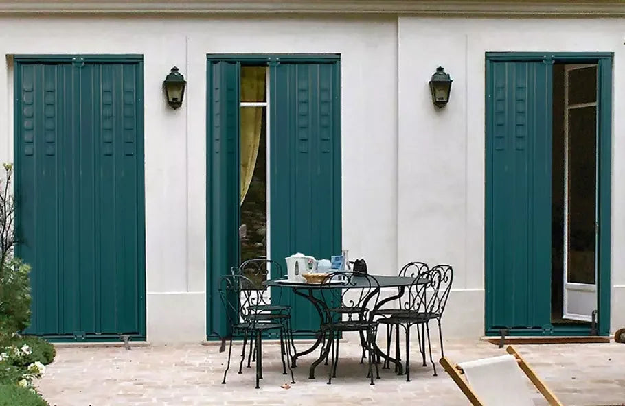 Terrasse avec table en fer forgé et chaises, devant une façade blanche équipée de persiennes vertes, luminaires muraux