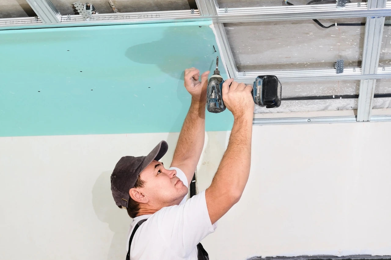 Plaquiste fixant une plaque de plâtre verte sur une ossature métallique de plafond à l’aide d’une visseuse.