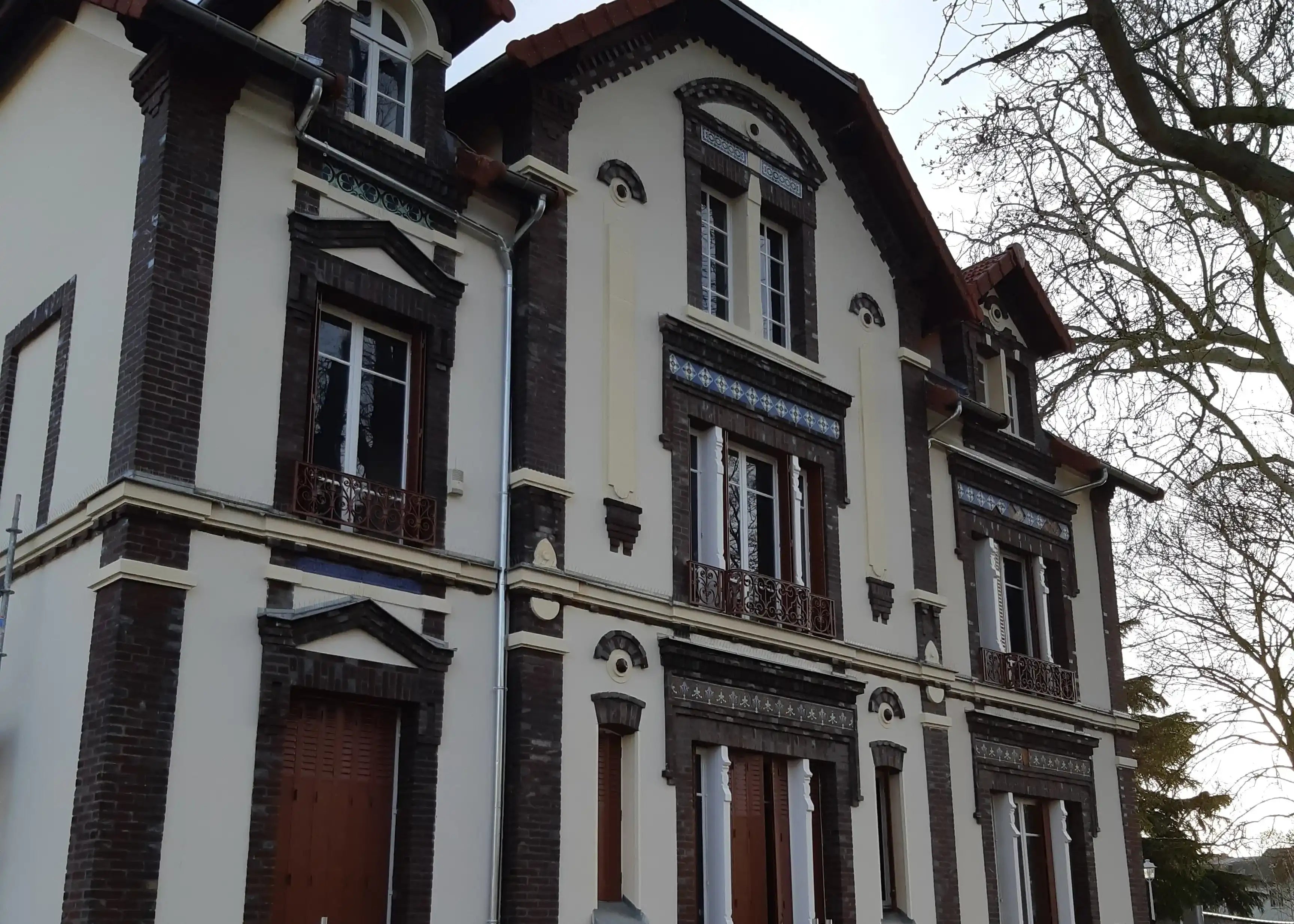 Façade rénovée en briques et enduit d'une maison ancienne avec détails décoratifs et volets en bois, sur fond d'arbre